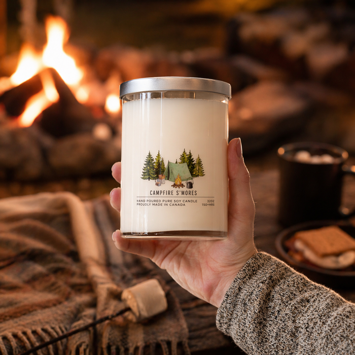 Hand holding a candle labeled 'Campfire S'mores' in front of a cozy fireplace.